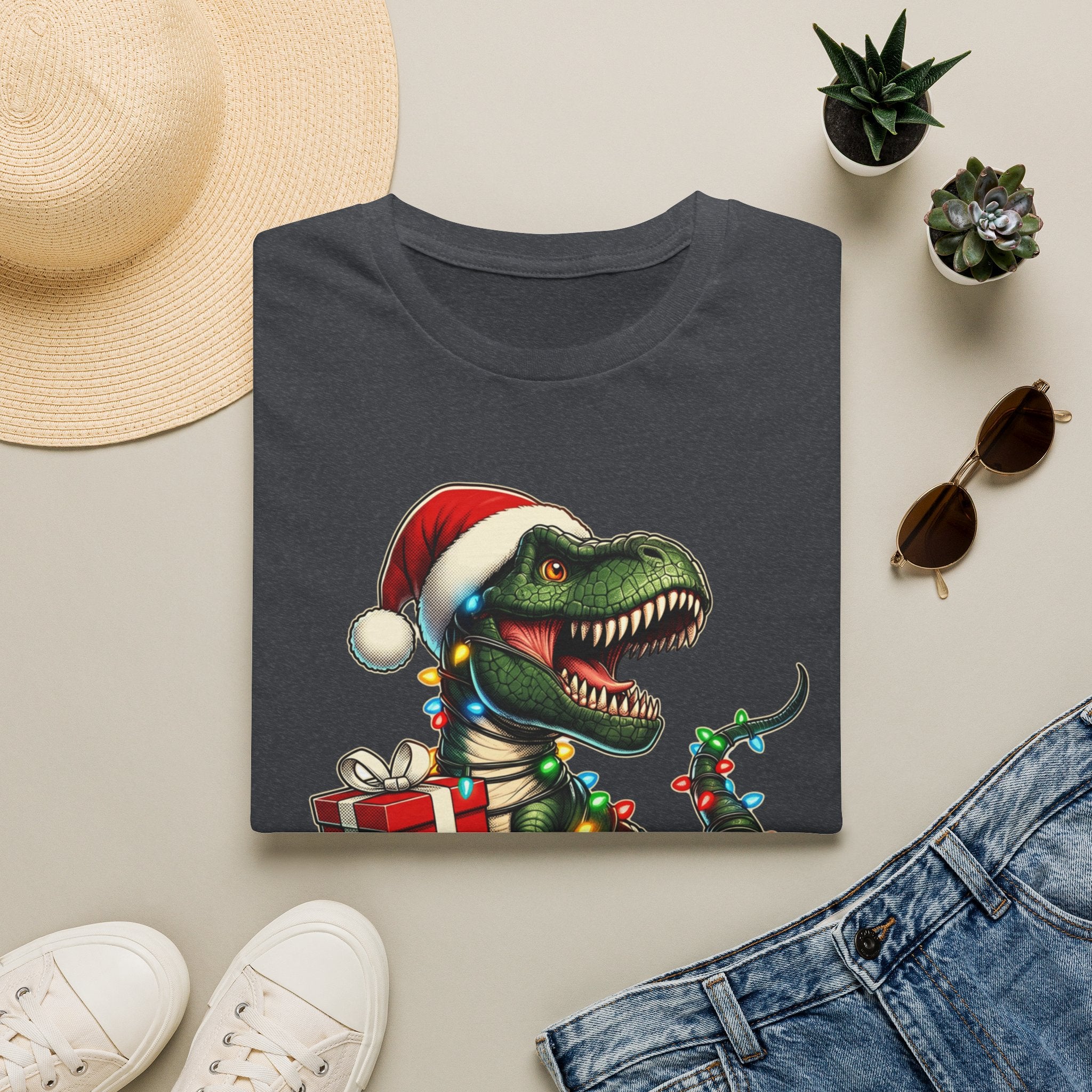 Happy Saurus T-Rex Santa Hat Christmas Lights print on a Dark Heather Grey t-shirt, folded beside a straw hat and jeans
