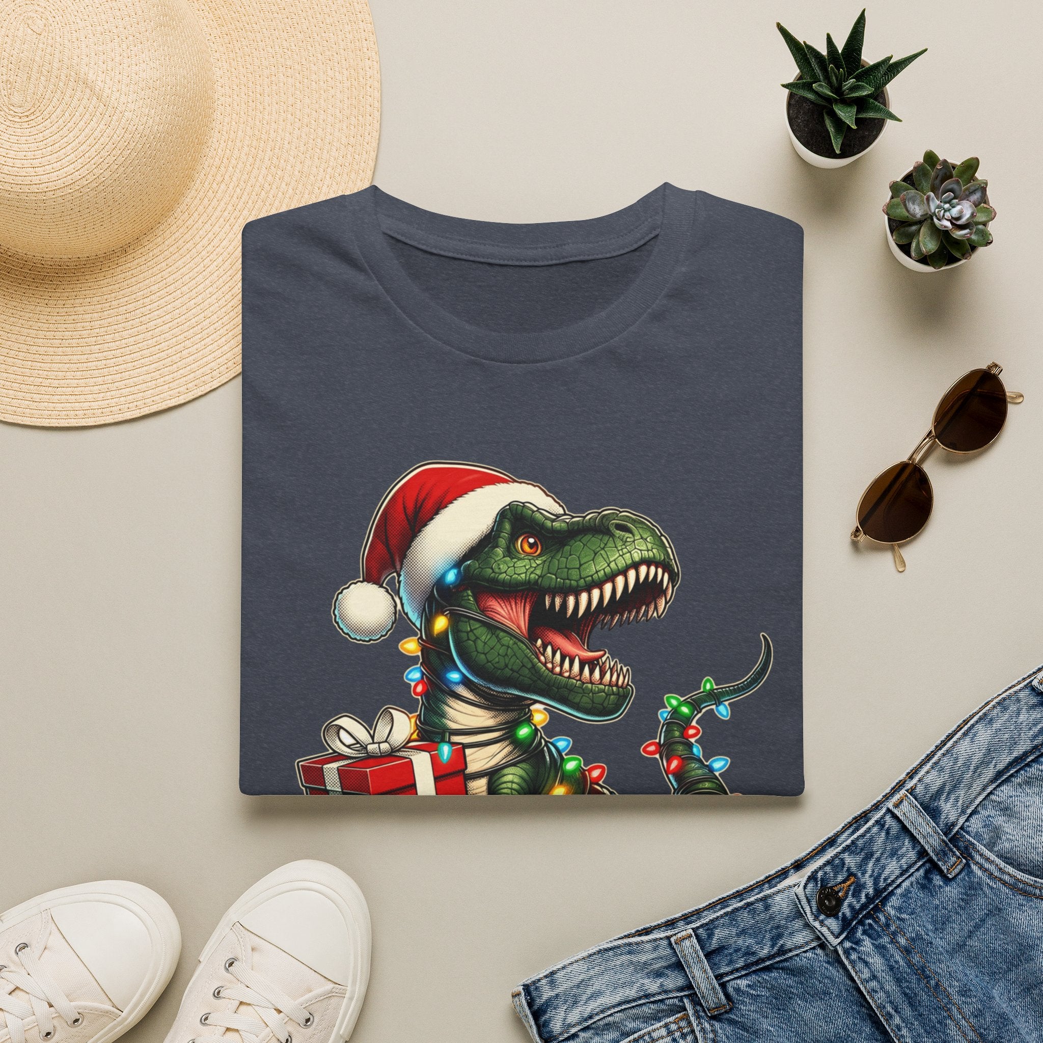 Happy Saurus T-Rex Santa Hat Christmas Lights print on a Heather Navy t-shirt, folded beside a straw hat and jeans