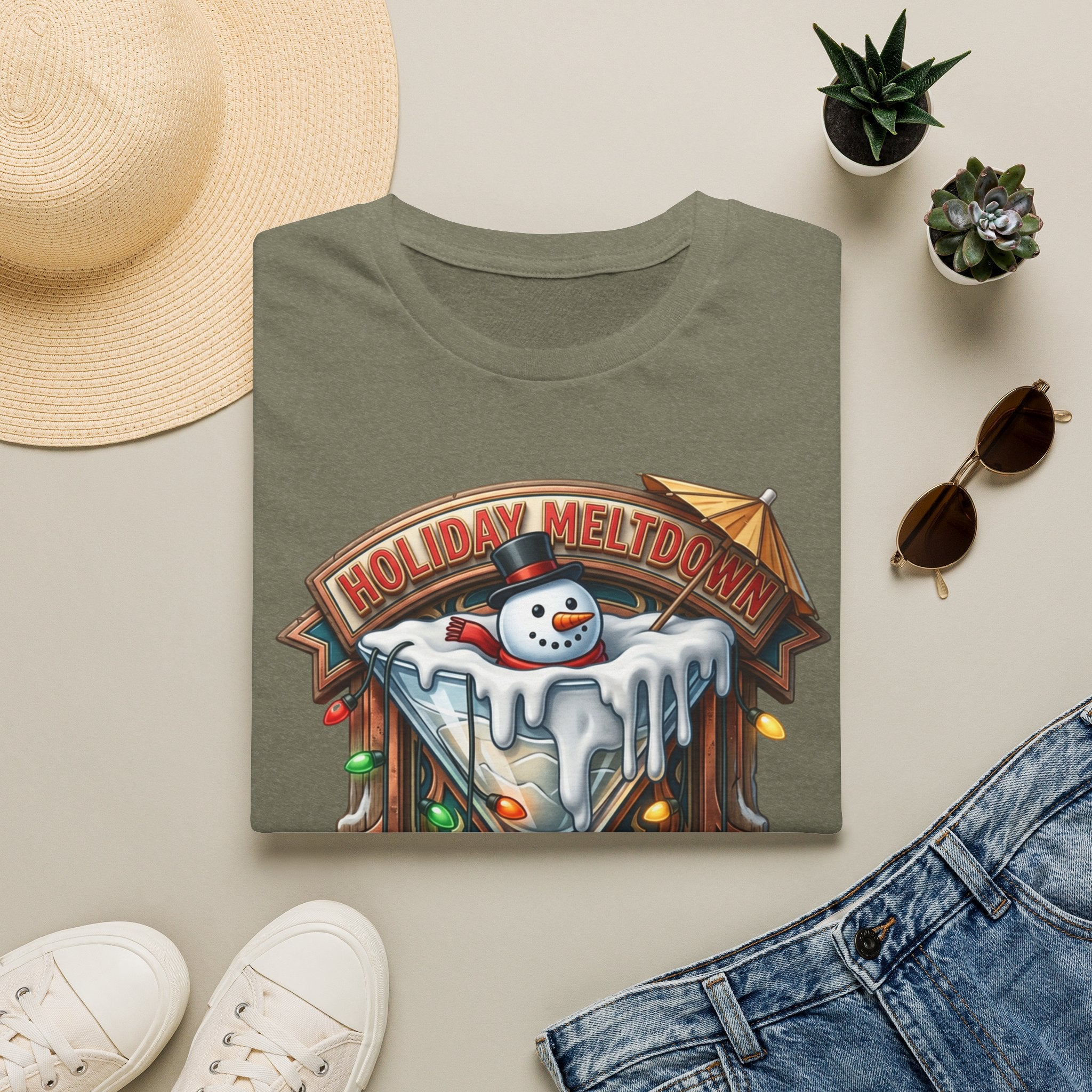 Holiday Meltdown Snowman Martini print on a Heather Military Green t-shirt, folded beside a straw hat and jeans