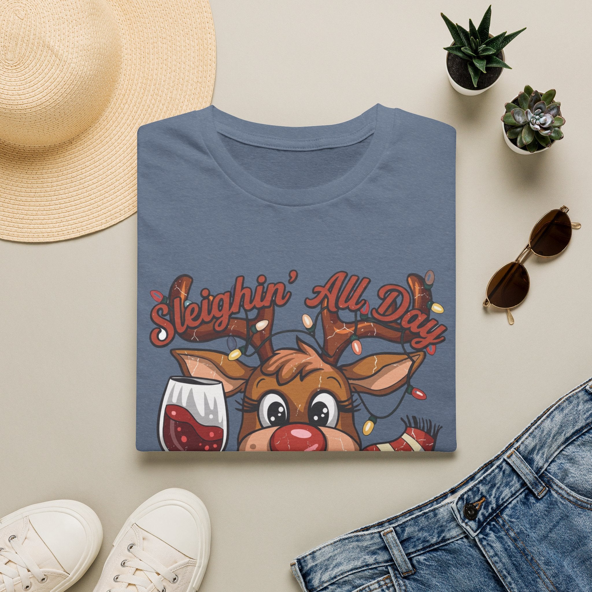 Reindeer Sleighin' All Day Tipsy All Night print on a Heather Indigo t-shirt, folded beside a straw hat and jeans
