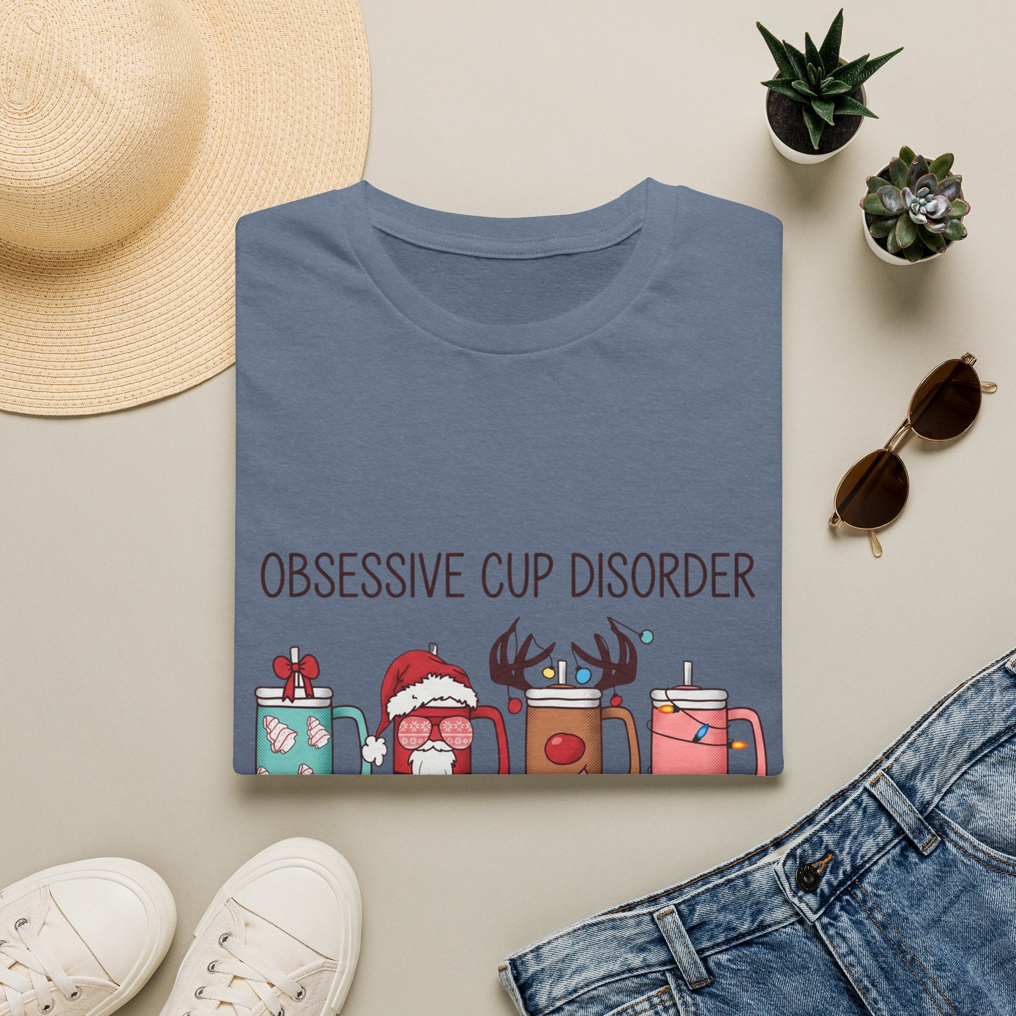 Obsessive Cup Disorder Funny Christmas Tumbler print on a Heather Indigo t-shirt, folded beside a straw hat and jeans