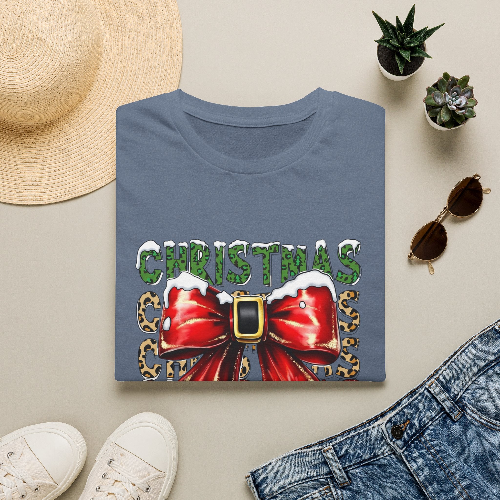 Stacked Christmas Leopard Print Santa Belt Bow print on a Heather Indigo t-shirt, folded beside a straw hat and jeans