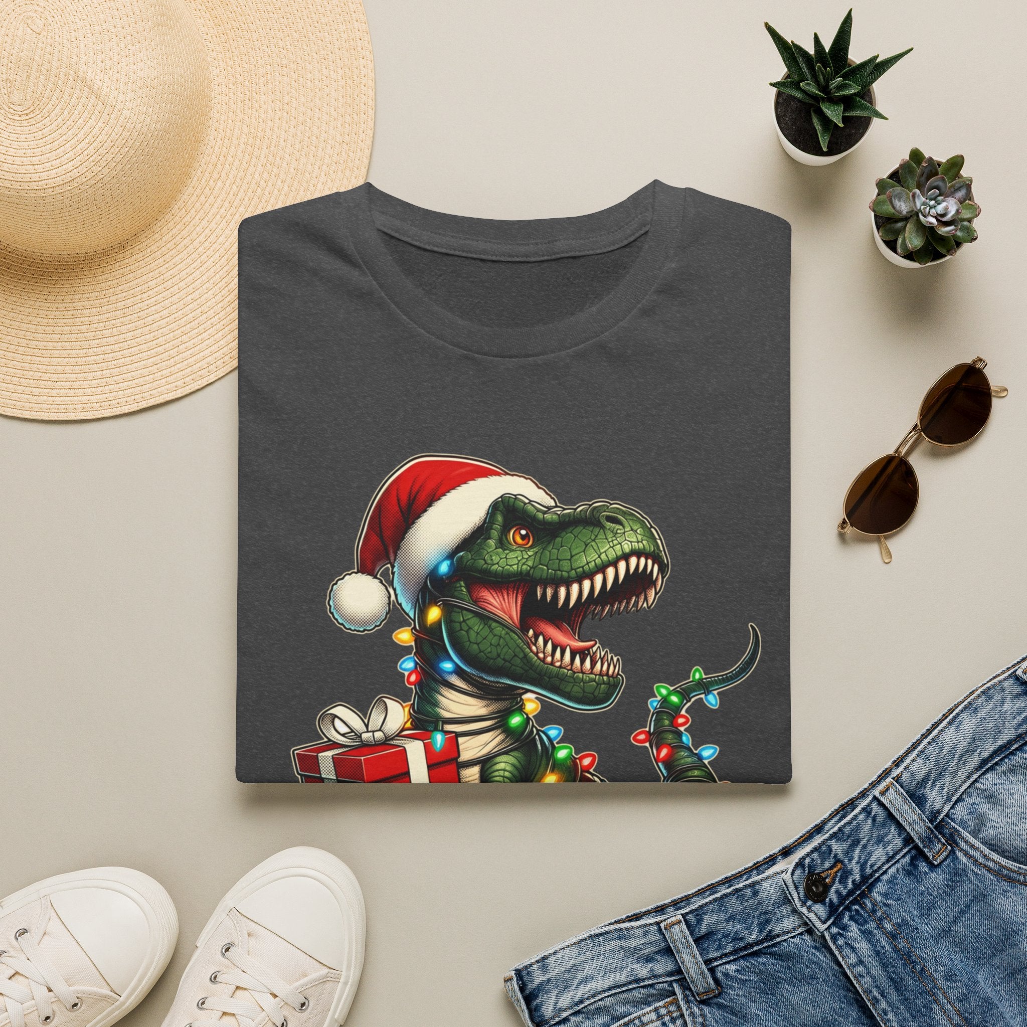 Happy Saurus T-Rex Santa Hat Christmas Lights print on a Dark Heather t-shirt, folded beside a straw hat and jeans