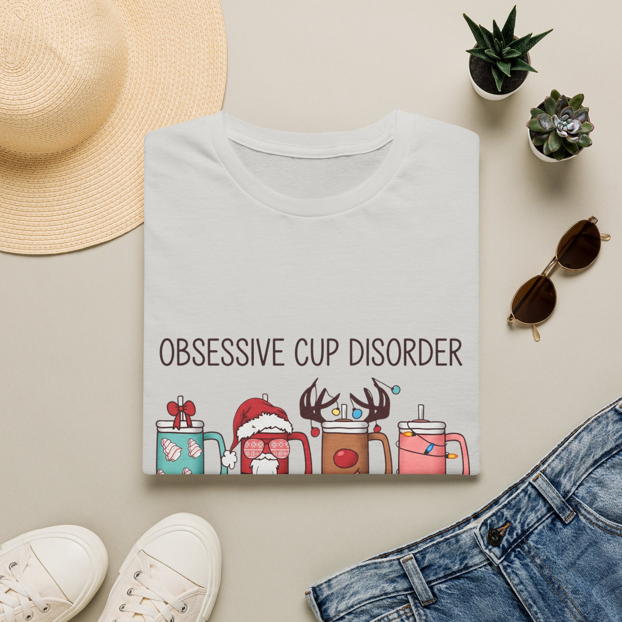 Obsessive Cup Disorder Funny Christmas Tumbler print on an Ice Grey t-shirt, folded beside a straw hat and jeans