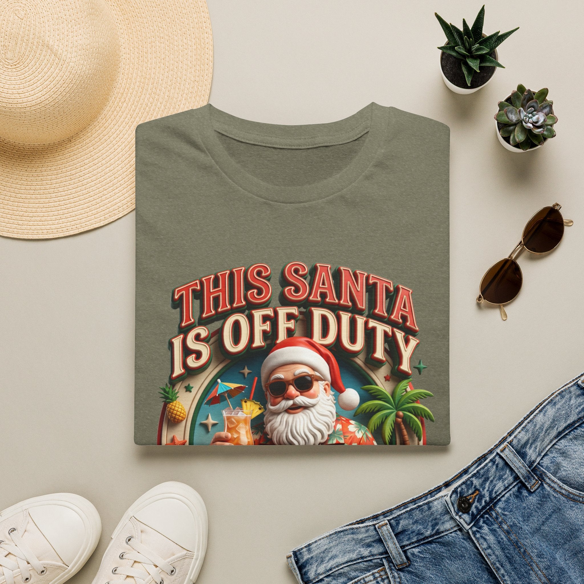 This Santa Is Off Duty Cocktail Club print on a Heather Military Green t-shirt, folded beside a straw hat and jeans