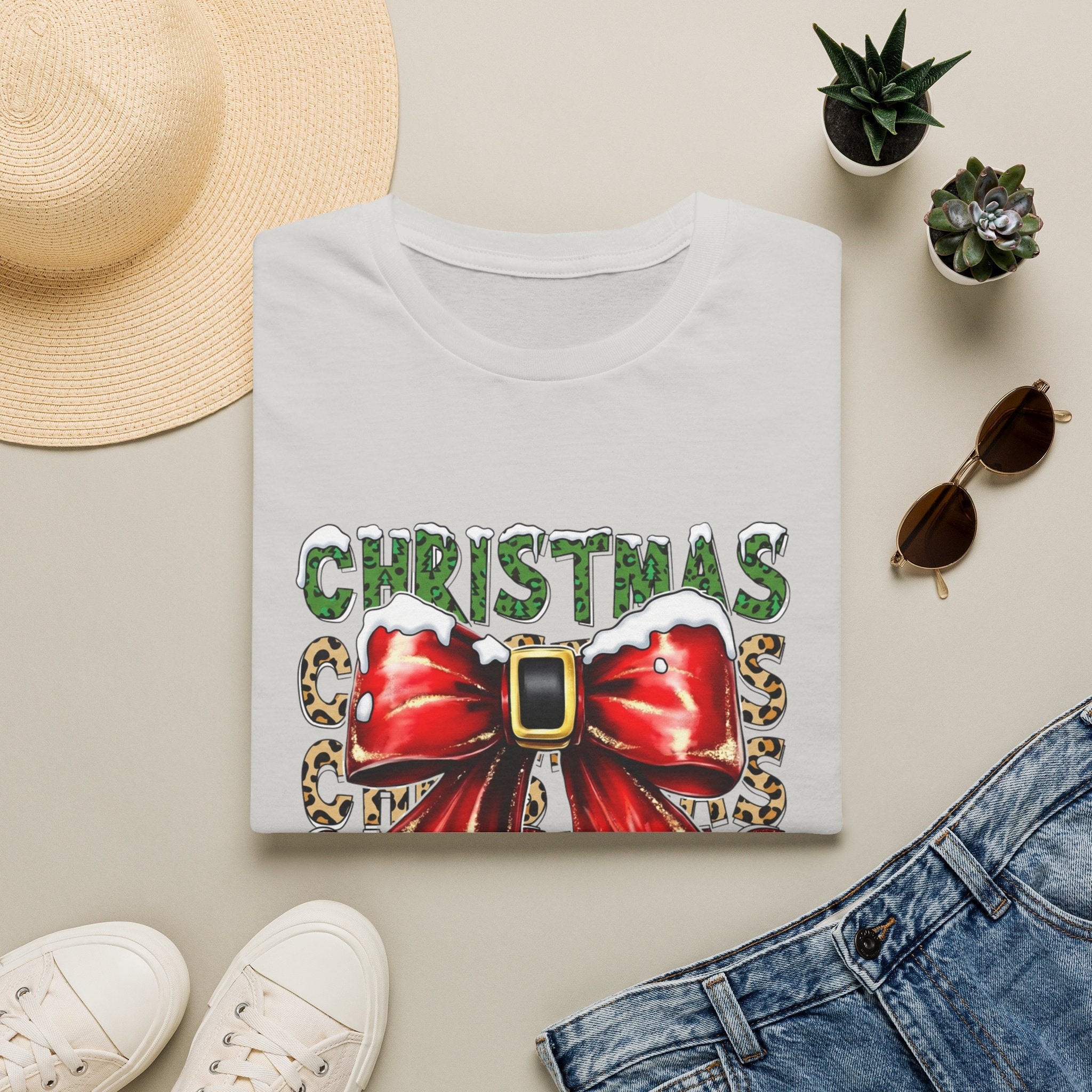 Stacked Christmas Leopard Print Santa Belt Bow print on an Ice Grey t-shirt, folded beside a straw hat and jeans