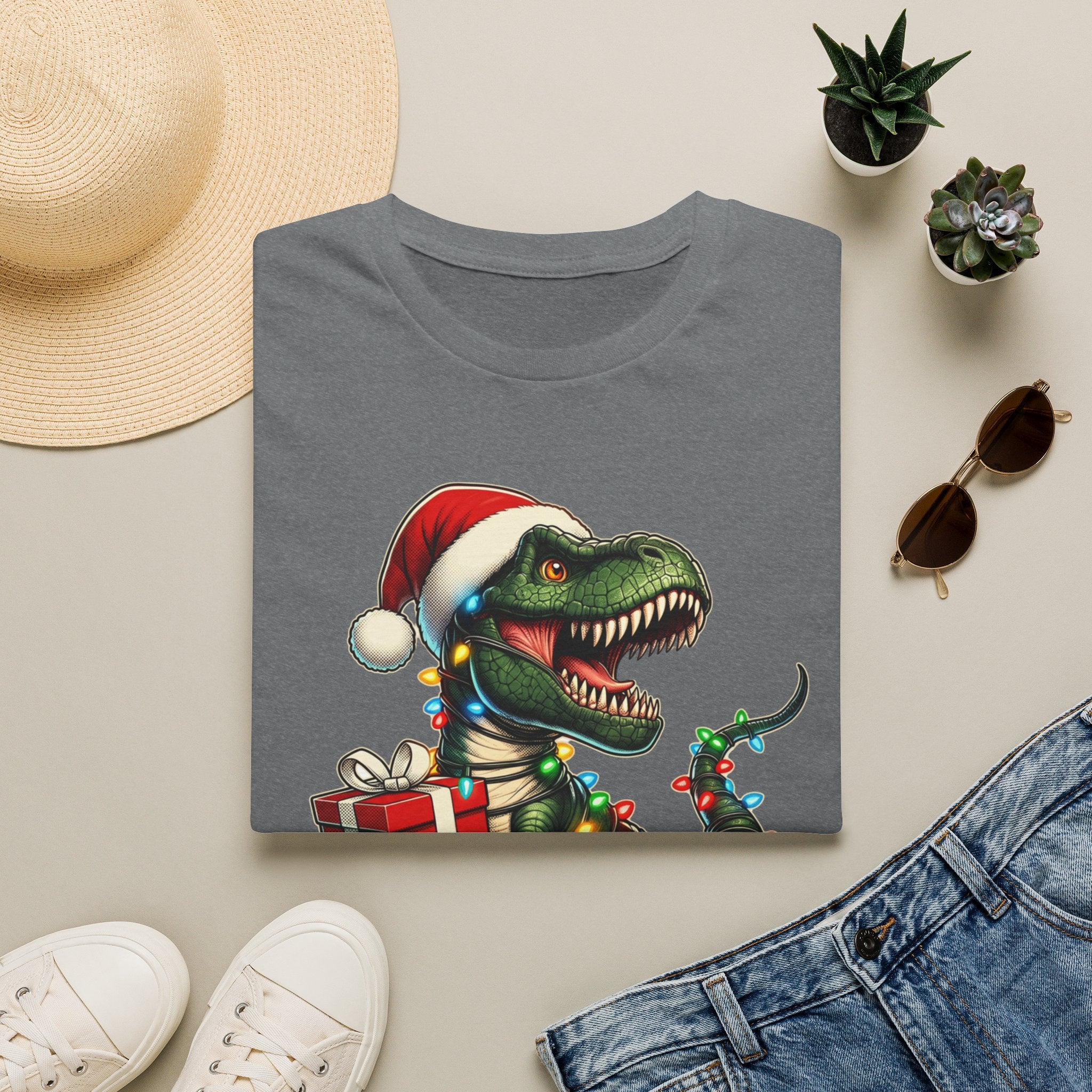 Happy Saurus T-Rex Santa Hat Christmas Lights print on a Graphite Heather t-shirt, folded beside a straw hat and jeans