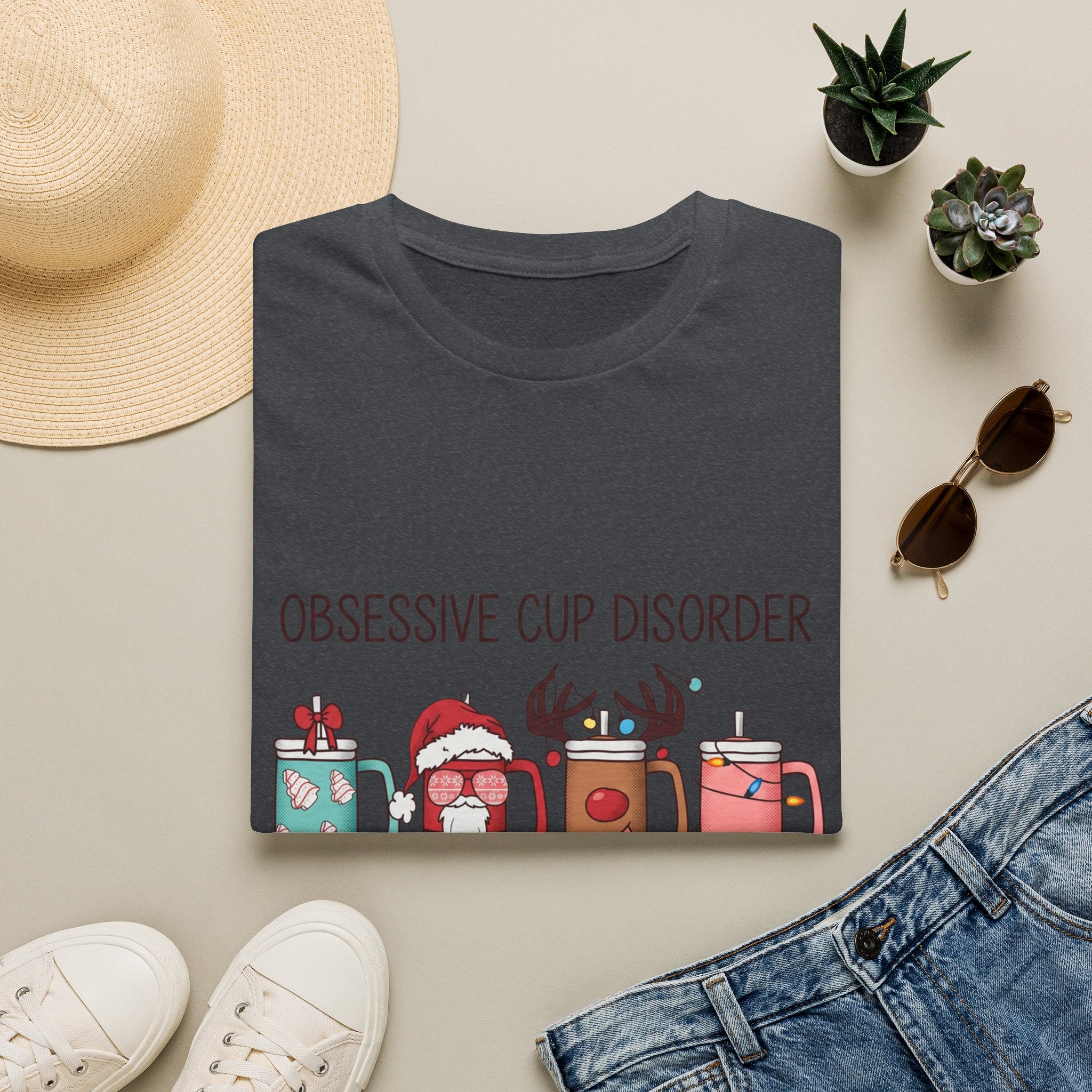 Obsessive Cup Disorder Funny Christmas Tumbler print on a Dark Heather Grey t-shirt, folded beside a straw hat and jeans