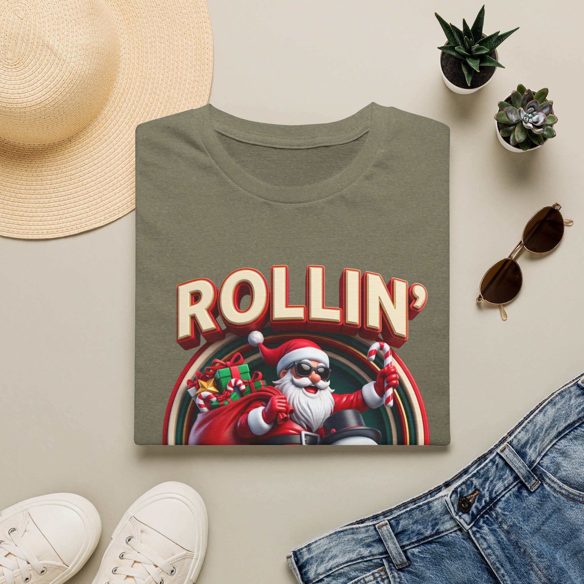 Rollin' with My Snowmies Santa & Snowman print on a Heather Military Green t-shirt, folded beside a straw hat and jeans