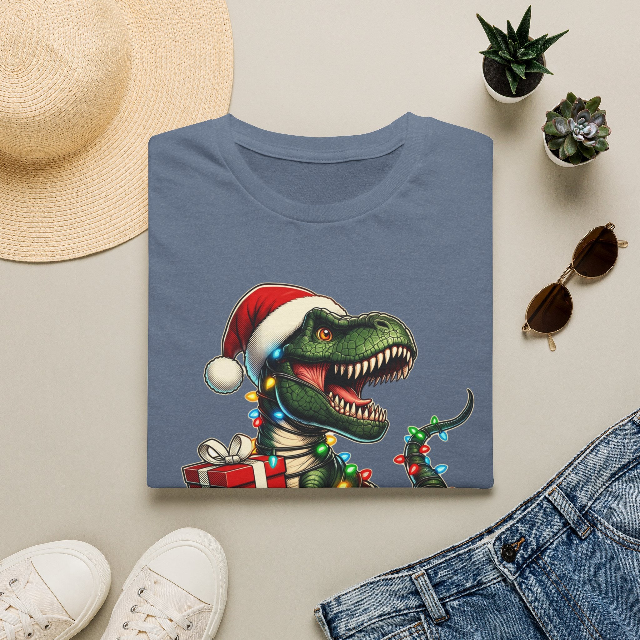 Happy Saurus T-Rex Santa Hat Christmas Lights print on a Heather Indigo t-shirt, folded beside a straw hat and jeans