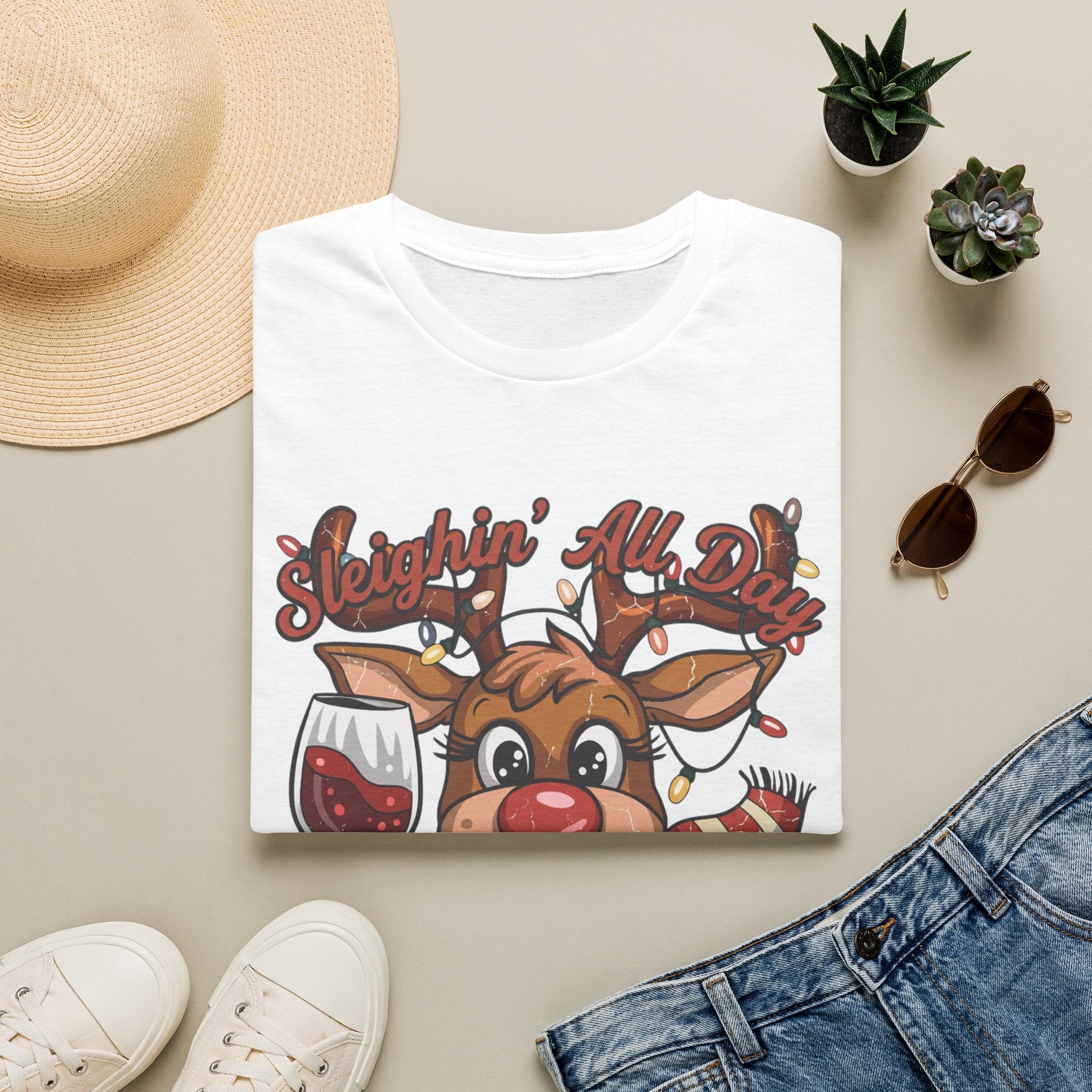 Reindeer Sleighin' All Day Tipsy All Night print on a White t-shirt, folded beside a straw hat and jeans