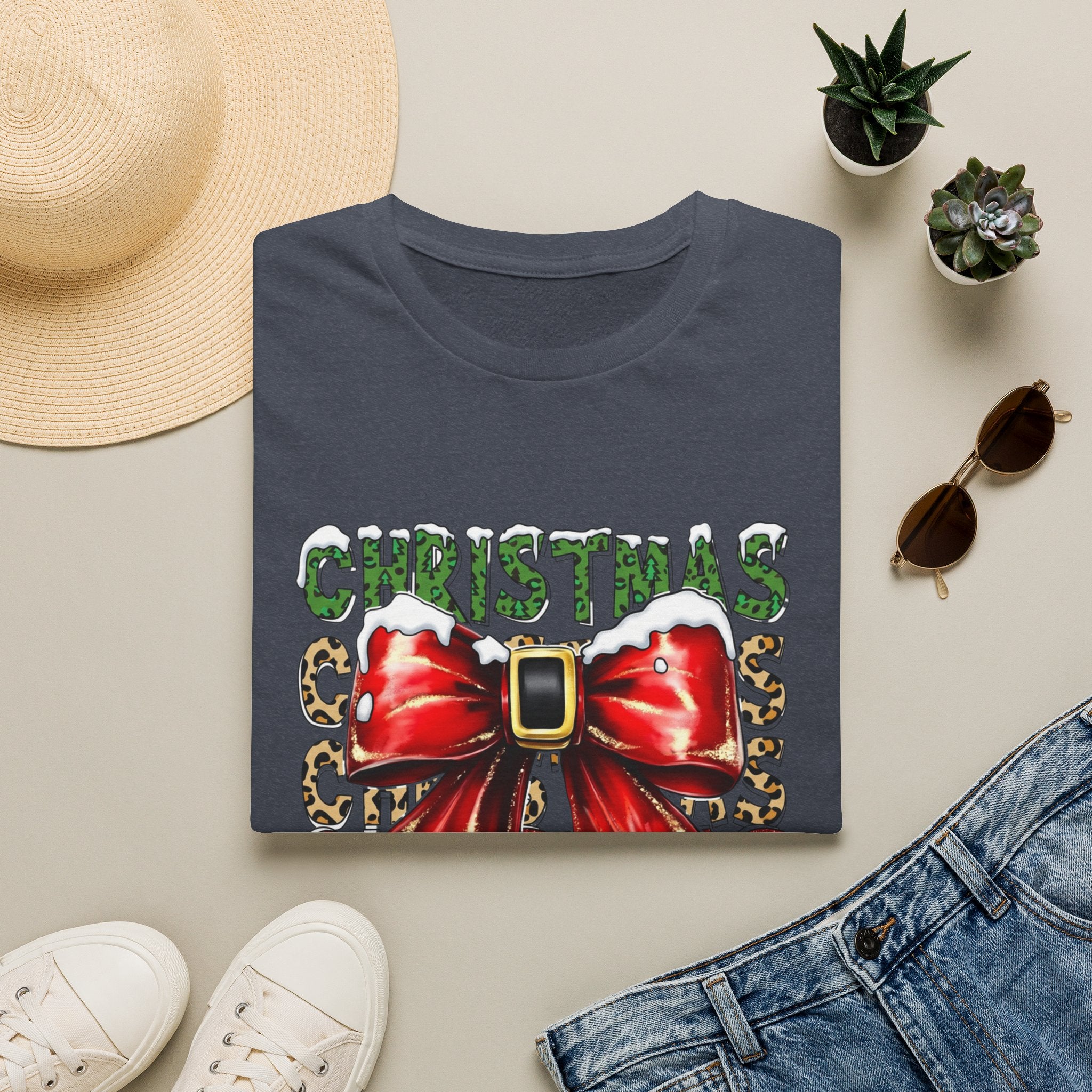 Stacked Christmas Leopard Print Santa Belt Bow print on a Heather Navy t-shirt, folded beside a straw hat and jeans