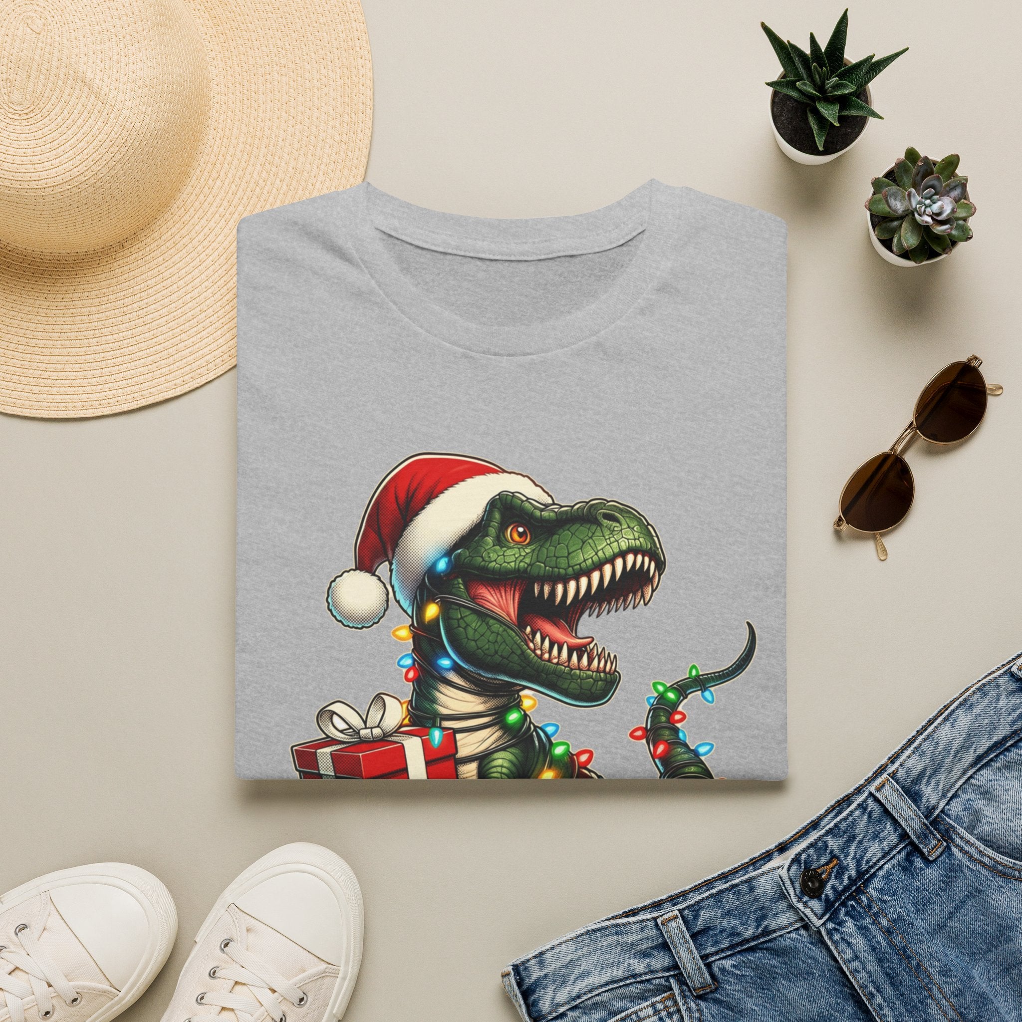 Happy Saurus T-Rex Santa Hat Christmas Lights print on a Sport Grey t-shirt, folded beside a straw hat and jeans