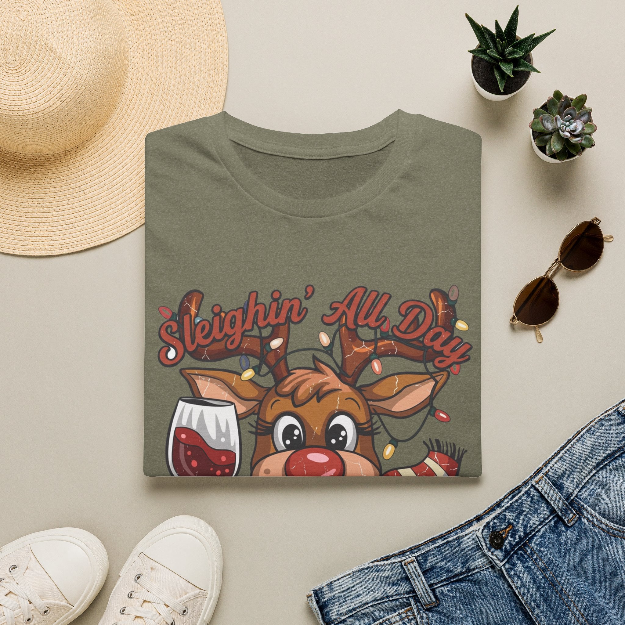 Reindeer Sleighin' All Day Tipsy All Night print on a Heather Military Green t-shirt, folded beside a straw hat and jeans