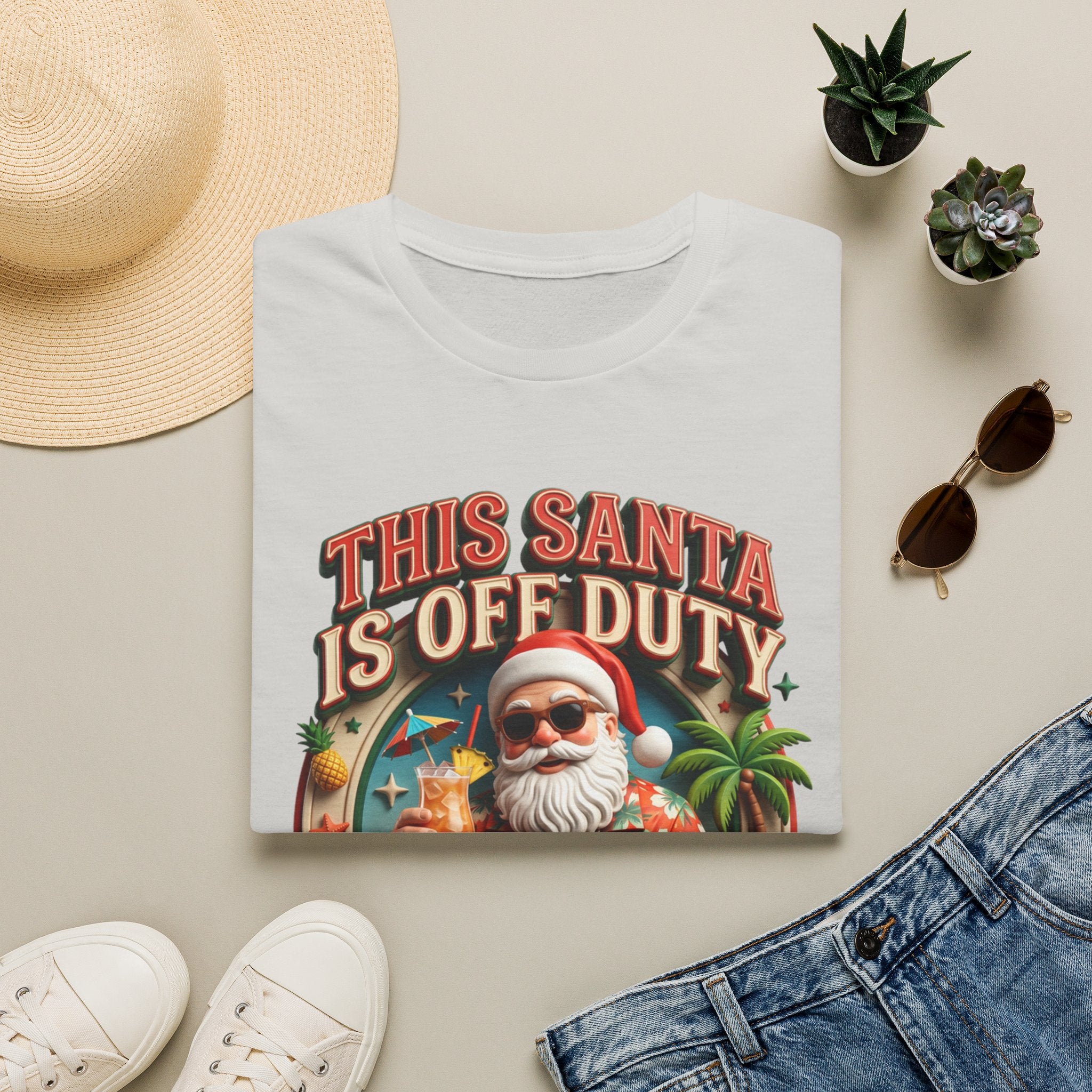 This Santa Is Off Duty Cocktail Club print on an Ice Grey t-shirt, folded beside a straw hat and jeans