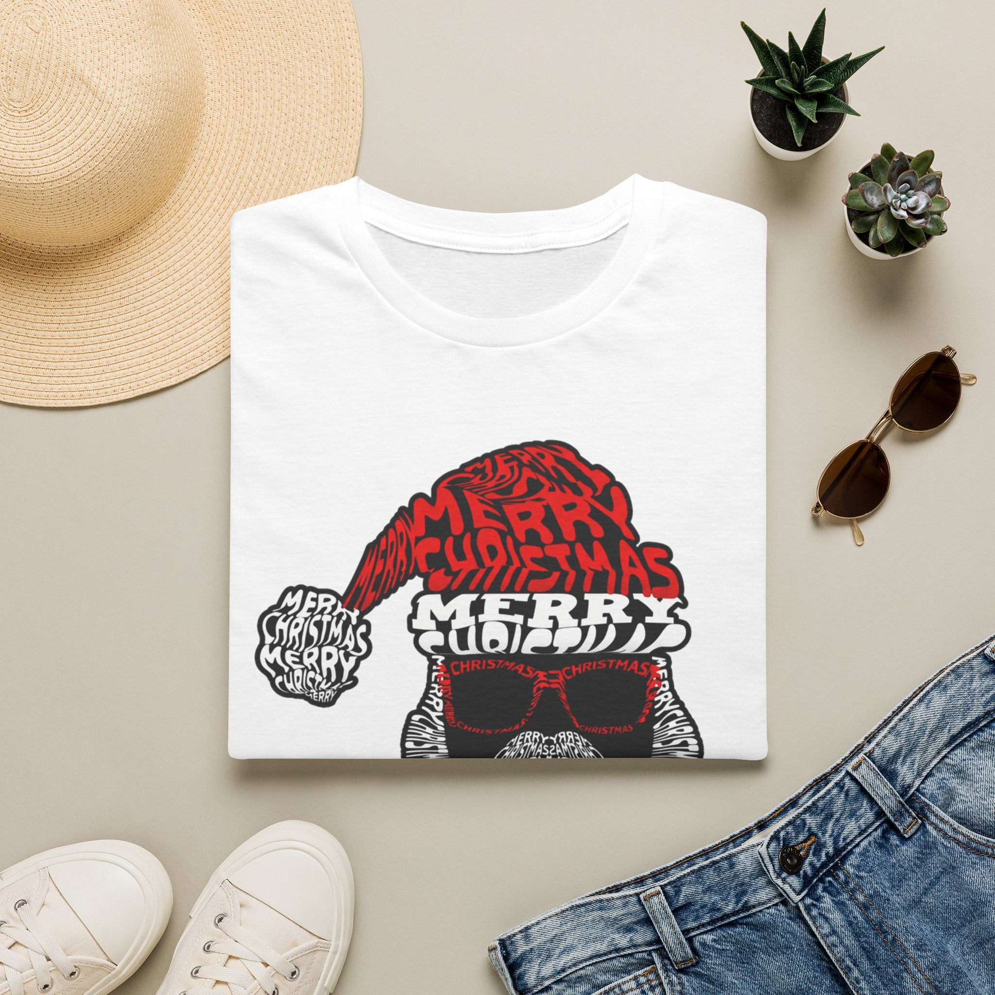 Cool Santa Sunglasses Typography Merry print on a White t-shirt, folded beside a straw hat and jeans