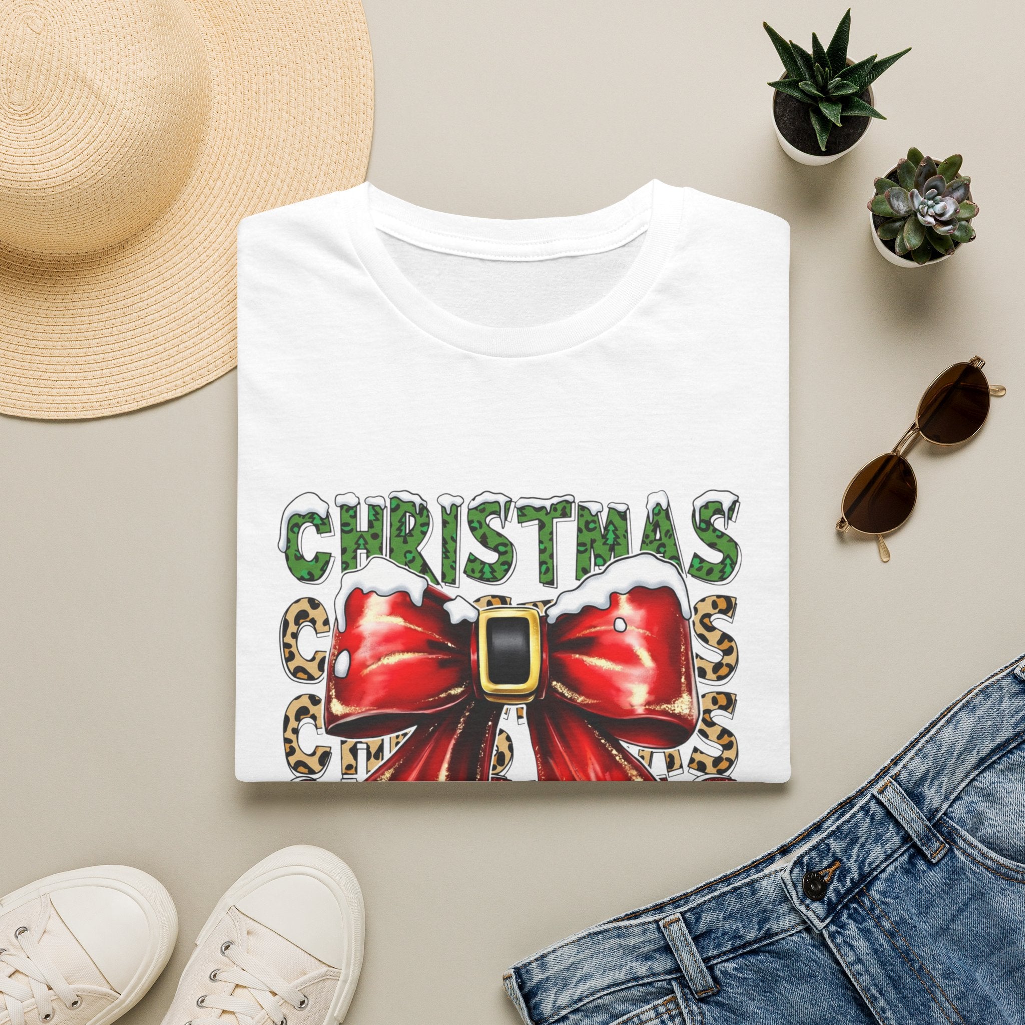 Stacked Christmas Leopard Print Santa Belt Bow print on a White t-shirt, folded beside a straw hat and jeans