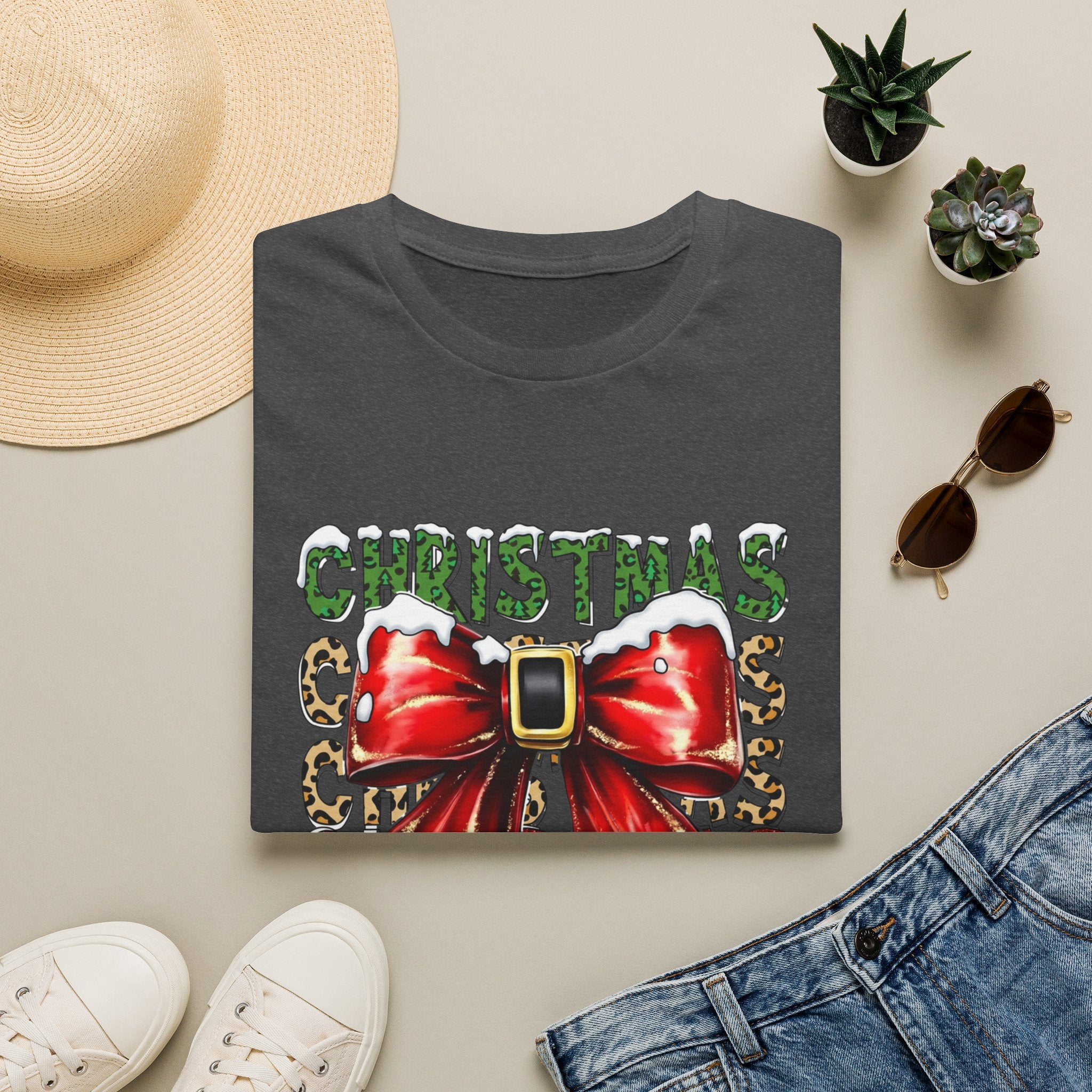 Stacked Christmas Leopard Print Santa Belt Bow print on a Dark Heather t-shirt, folded beside a straw hat and jeans