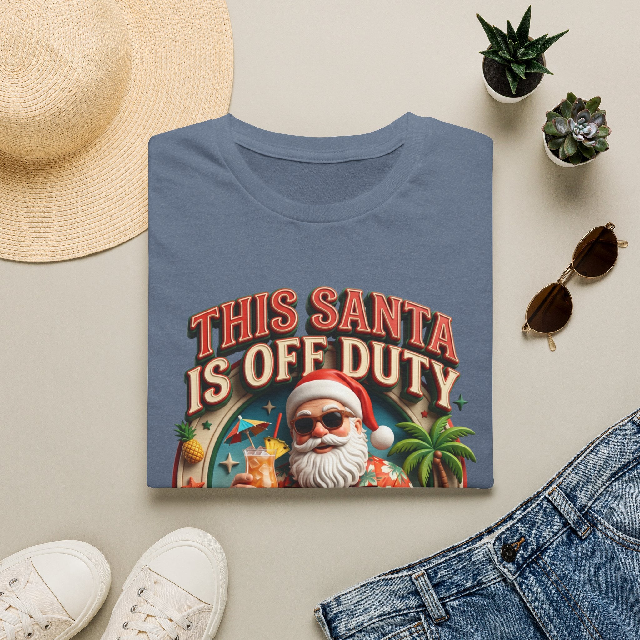 This Santa Is Off Duty Cocktail Club print on a Heather Indigo t-shirt, folded beside a straw hat and jeans