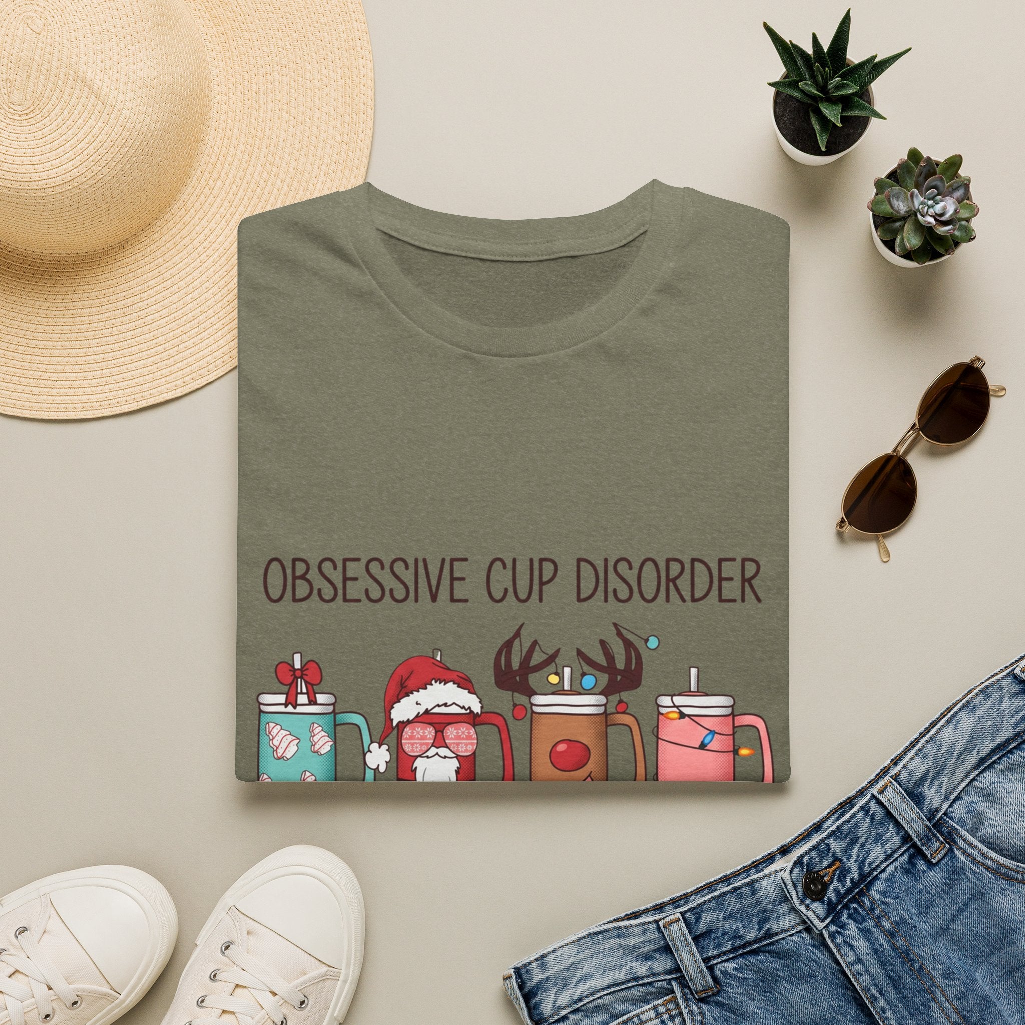Obsessive Cup Disorder Funny Christmas Tumbler print on a Heather Military Green t-shirt, folded beside a straw hat and jeans
